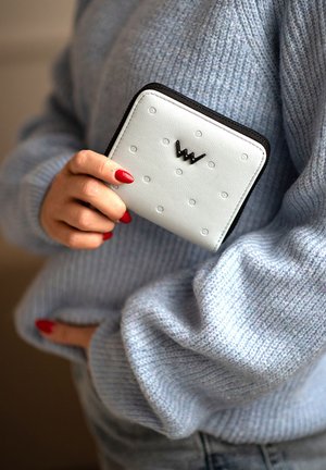 Light blue leather wallet with embossed dots, black zip, and a distinctive black logo. The wallet is held in a hand with red nails.