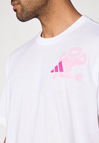 Biele bavlnené tričko s jemne ružovou grafikou svalnatého charakteru držíaceho činky a štyrmi ružovými pruhmi na prednej strane.