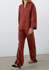 Haut en lin couleur rouille avec un col en V et des manches larges, associé à un pantalon assorti. Le pantalon est ample et descend jusqu'aux pieds.