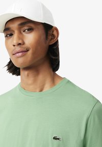 Groene katoen T-shirt met een ronde halslijn en een geborduurd logo op de borst; gedragen met een witte pet met een logo aan de voorkant.