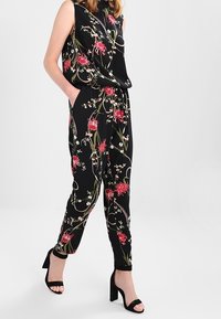 Svart blommig jumpsuit med röda och vita mönster, ärmlös design, sidofickor och tapered ben. Matchas med svarta högklackade sandaler.