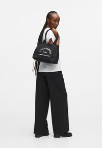 Melnas auduma iepirkumu soma ar baltu tekstu, kas lasa "KARL LAGERFELD PARIS 21 RUE ST-GUILLAUME", ar izturīgām rokturiem un kvadrātveida formu.