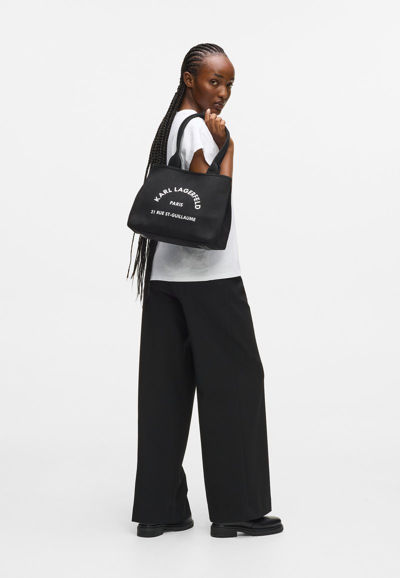 Melnas auduma iepirkumu soma ar baltu tekstu, kas lasa "KARL LAGERFELD PARIS 21 RUE ST-GUILLAUME", ar izturīgām rokturiem un kvadrātveida formu.
