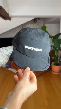 Kéz tart egy sötétszürke Dickies sapkát beltéren, háttérben fa padló és zöld cserepes növény látható.