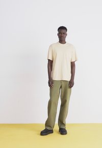 Tricou din bumbac bej cu un buzunar, asortat cu pantaloni verzi măslinii și saboți negri, stând pe un podea galbenă, în fața unui perete alb.
