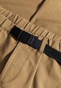 Khaki spodnie z marszczonym pasem, z czarnym paskiem z plastikową klamrą. Gładka faktura tkaniny i dwie widoczne kieszenie boczne.