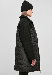 Čierny prešívaný kabát s plyšovými, textúrovanými rukávmi a golierom. Má zips a bočné vrecká. Nosený s čiernym beanie.