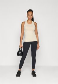 Beige αμάνικο τοπ συνδυασμένο με μαύρα κολάν. Το μοντέλο κρατάει μια μαύρη αλτήρα kettlebell, με υφαντό ύφασμα και εφαρμοστή σχεδίαση.