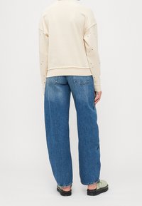 Personne portant un haut à manches longues beige clair avec de petits points noirs et un jean bleu ample, vue de dos sur un fond uni.
