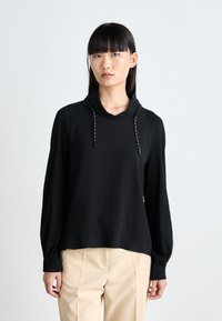 Black hooded top with long sleeves, featuring drawstrings and a relaxed fit, paired with beige trousers, against a neutral background.