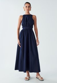 Kvinna som står i en ärmlös, marinblå maxiklänning med insydd midja, bärande vita sandaler med remmar och guldörhängen i form avringar.