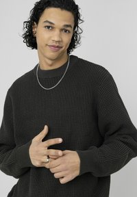 Pull texturé vert foncé avec col rond, poignets côtelés, et motif subtil. Le mannequin porte un collier en chaîne argentée et une bague.