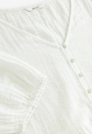 Blusa bianca a maniche corte con scollo a V, caratterizzata da chiusure con bottoni e un tessuto testurizzato con piccoli punti in rilievo. Materiale morbido e leggero.