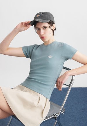 Noor istub metalltoolil, kandes sinakas-halli kitsast T-särki, beežikat seelikut ja tumedat halli nokamütsi, hoides ühe käega mütsi ääre peal.