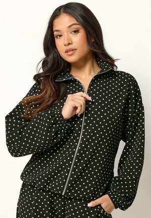 Vrouw met lang donker haar, gekleed in een zwart jasje met rits en bijpassende broek met witte stippen, poseert met één hand dicht bij de rits.