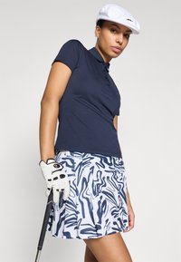 Jūras zila polo krekls ar īsām piedurknēm, kombinēts ar baltiem un jūras zila rakstainiem svārkiem ar krokām. Uzvilkta balta cepure un balta golfa cimdi.