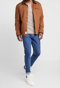 Brun canvasjacka med mörk krage, beige tröja, blå jeans och vita sneakers. Jackan har knappar och en bröstficka.