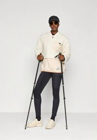 Šviesios spalvos striukė su priekinė kišene, derinama su tamsiai mėlynomis tamprėmis ir smėlio spalvos bateliais. Modelis laiko žygiavimo lazdas, dėvi saulės akinius ir kepurę.