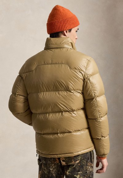 Personne portant une doudoune marron clair, un bonnet en tricot orange et un pantalon camouflage, vue de dos sur un fond neutre.
