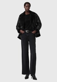 Czarna skórzana kurtka z podszewką z owczej wełny, czarny sweter, czarne spodnie z wysokim stanem i czarne botki na obcasie. Modelka stoi z rękami na biodrach.