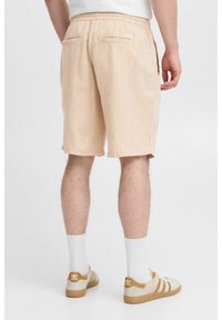 Beige shorts, valmistettu tekstuuripinnasta, rentoa mallia, jossa on joustava vyötärö ja kaksi takataskua. Yhdistetty valkoisten sukkien ja beigejen lenkkarien kanssa.