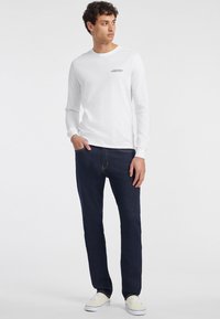 Balta ilgomis rankovėmis marškiniai su juodu "GUESS JEANS" užrašu, derinami su tamsiai mėlynomis džinsomis ir šviesiais sportbačiais.