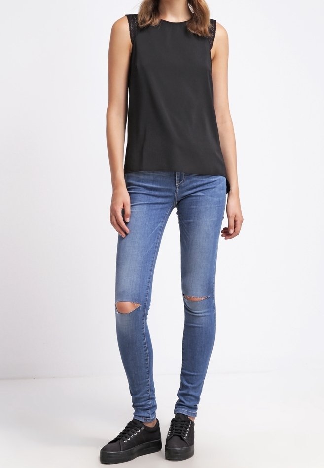 Schwarzes ärmelloses Oberteil mit Spitze, blaue Skinny-Jeans mit Knieflicken und schwarze Sneakers. Der Stoff ist glatt und hat ein figurbetontes Design.
