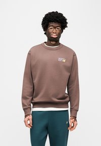 Καφέ βαμβακερό φούτερ με ριμπ μανσέτες και λαιμόκοψη, με μικρό λογότυπο New Balance. Συνδυασμένο με σκούρο πράσινο-μπλε φόρμα παντελόνι.