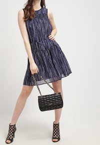 Dunkelblaues ärmelloses Kleid mit vertikalen weißen Mustern, schwingendem Rock; schwarze gesteppte Handtasche mit Kettenriemen; schwarze hochhackige Sandalen mit Netzdesign.