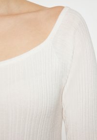 Top bianco a costine con scollatura ampia e maniche lunghe. Il tessuto ha una texture morbida con delicate linee verticali.