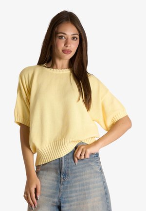 Donna con lunghi capelli castani che indossa un maglione giallo chiaro a maniche corte e jeans blu, posando con una mano sui fianchi.