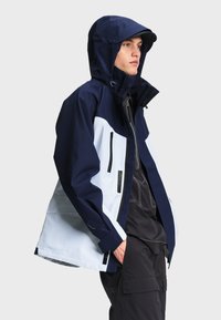 Jeune homme portant une veste à capuche bleu marine et blanche avec des poches zippées, un pantalon cargo noir et une casquette noire, regardant sur le côté.