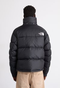 Svart puffjacka med hög krage, quiltad design och en logotyp på vänster axel. Slät tyg med en glänsande finish.