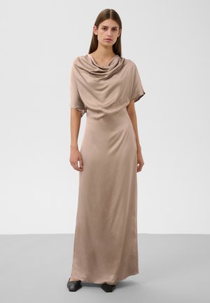 Vrouw draagt een lange beige satijnen jurk met een geplooide halslijn en korte mouwen, staand op een effen grijze vloer met een witte achtergrond.