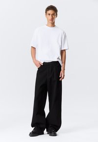 T-shirt blanc à manches courtes et pantalon noir ample en coton, avec une fermeture éclair et un design classique à 5 poches. Chaussures noires.