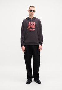 Mørkegrå hettegenser med en rød grafisk logo, kombinert med svarte cargo-bukser med sidelommer. Modellen har på seg svarte solbriller og sko.