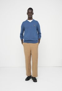 Blå v-ringad tröja, vit undertröja, beigefärgade byxor, svarta skor. Tröjan har en liten gul logotyp på bröstet. Slät textur.