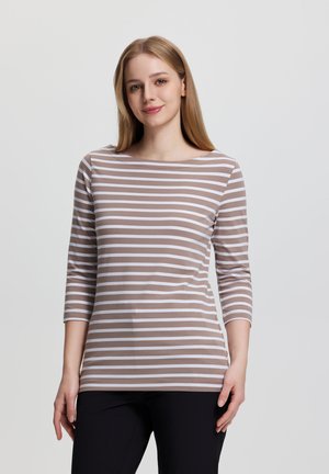 Mujer joven con cabello rubio lacio, vestida con una blusa de manga larga a rayas beige y blanca y pantalones negros, de pie frente a un fondo liso.