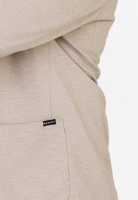 Beige textielkleding met een zichtbare naad en een klein zwart label met de tekst "KLEINIGKEIT" nabij de rand.