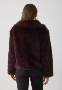 Bordeaux nep-bont jas met een hoge kraag, lange mouwen en een zachte, fluffy textuur; gecombineerd met zwarte jeans.