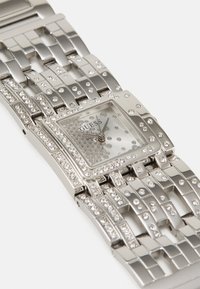 Montre en métal argenté avec un boîtier rectangulaire, orné de strass, dotée d'un cadran transparent et d'un bracelet métallique à maillons.