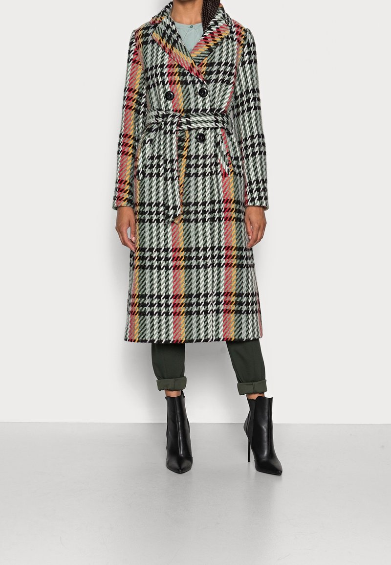Femme portant un long manteau en laine ceinturé avec un grand motif à carreaux noir, blanc, rouge et jaune, associé à des bottines noires et un pantalon retroussé.