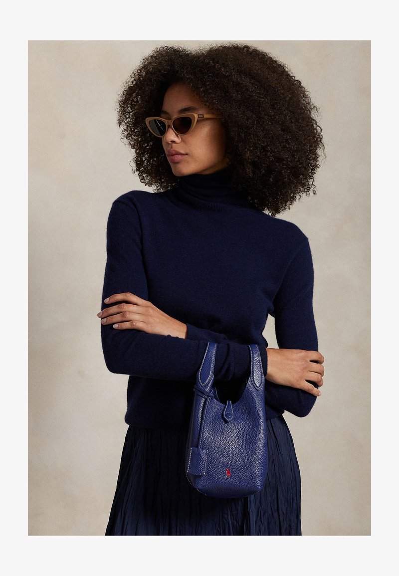 Woman with curly hair wearing brown cat-eye sunglasses and navy turtleneck holds small blue leather handbag against beige background.