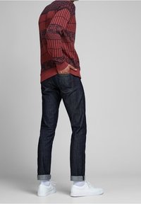 Roter und marineblauer gemusterter Strickpullover kombiniert mit dunklen Jeans, aufgerollten Bündchen und weißen Turnschuhen. Glatte Textur und figurbetontes Design.
