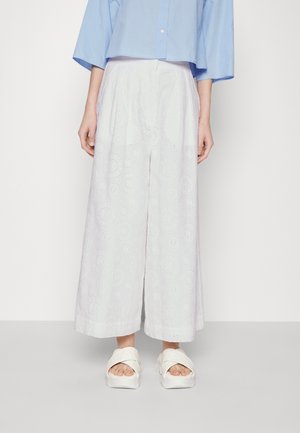 Žena v bílých širokých kalhotách s výšivkou, světle modré krátké blůze a bílých platformových sandálech, stojící před jednoduchým pozadím.