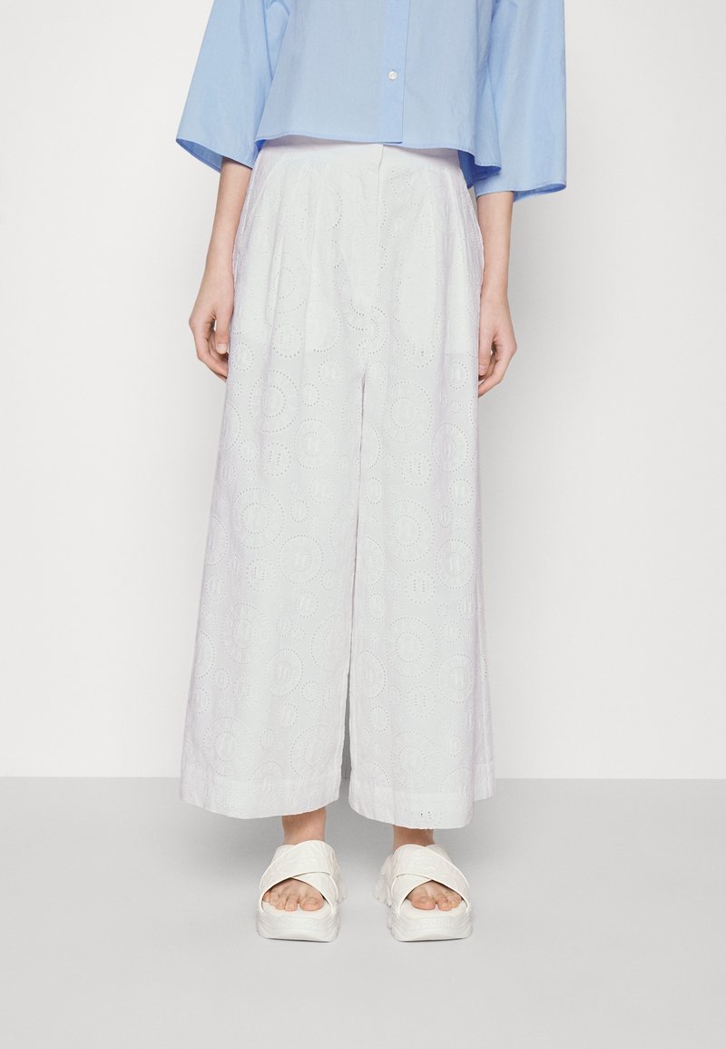 Woman wearing white wide-leg eyelet pants, light blue cropped blouse, and white platform sandals, standing against a plain background.