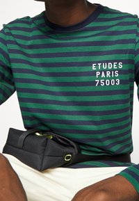 Gestreiftes langärmeliges Hemd in Dunkelgrün und Marineblau, mit weißem Text "ETUDES PARIS 75003". Schwarze Gürteltasche mit grünen Akzenten.
