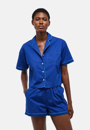 Femme portant une chemise bleue à manches courtes boutonnée et un short assorti avec un bord festonné bleu clair, debout devant un fond uni.