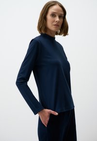 Marineblauwe lange mouw top met een hoge halslijn, ontspannen pasvorm, zachte textuur en minimale naden. Model poseert met één hand in een zijzak.