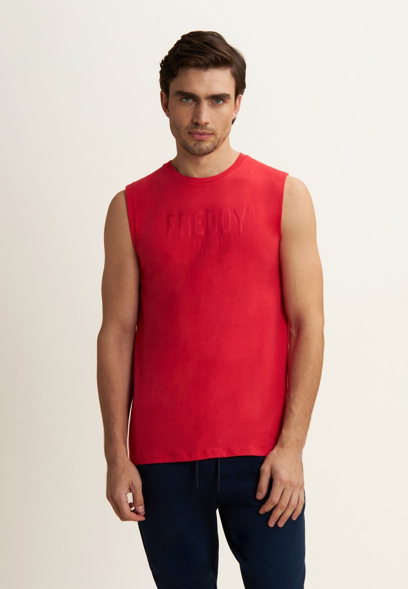 Hombre con cabello corto castaño, vistiendo una camiseta roja sin mangas y pantalones azul oscuro, de pie contra un fondo claro y liso.